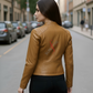 Woman wearing a tan leather jacket with a brand logo on a city street.