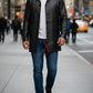 Man wearing a black leather coat and blue jeans walking on a city street.