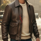Man wearing a brown leather jacket over a beige sweater with a red logo on a city street.